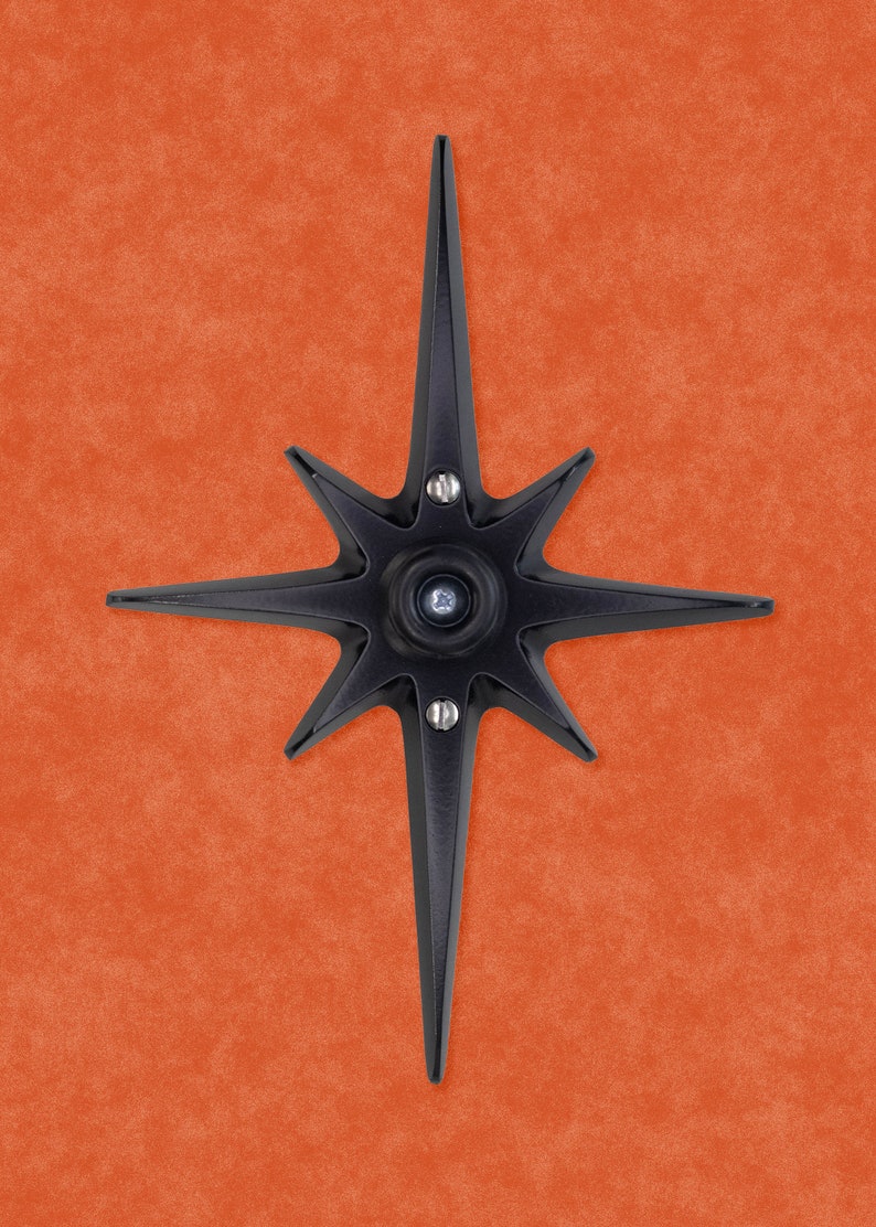 May include: A black metal starburst with eight points, each point ending in a sharp tip. The starburst has a circular center with a small hole in the middle.