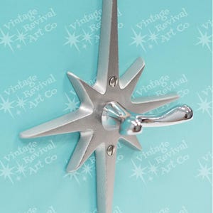 May include: A silver-toned metal wall hook featuring a starburst design with eight pointed rays. The center of the starburst has a smooth, curved hook extending outward, secured to the base with two visible flat-head screws in a vertical line.
