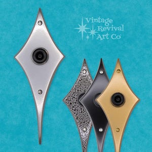 May include: A collection of diamond-shaped doorbells in silver, speckled, black, and gold finishes. Each doorbell has a central black button and is mounted with small silver screws. The background is a teal color, and the text "Vintage Revival Art Co" is visible.