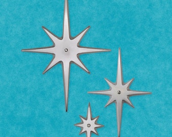May include: Three silver starburst wall decorations on a blue background. The stars are different sizes and have a pointed, six-pointed star shape.