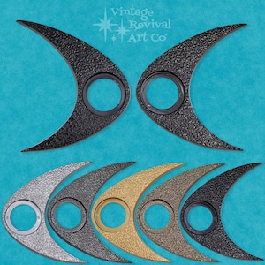 May include: Set of eight metal wall decor pieces in various finishes, including black, silver, gold, and bronze. Each piece features a crescent moon shape with a circular cutout in the center. The pieces are arranged in two rows, with the top row featuring two black pieces and the bottom row featuring six pieces in various finishes. The text "Vintage Revival Art Co." is visible in the top left corner of the image.