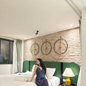 May include: A woman in a blue and purple dress sits on a white bed with a green headboard. The headboard has a brick wall behind it with three black and gold circular wall decorations. A lamp sits on a wooden nightstand next to the bed.