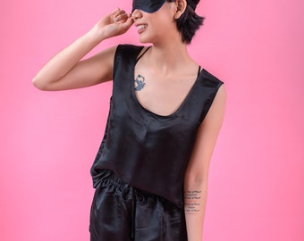 Puede incluir: Una mujer con una máscara de dormir negra, un top de satén negro y pantalones cortos de satén negro. Está sonriendo y mirando hacia un lado. El fondo es rosa.