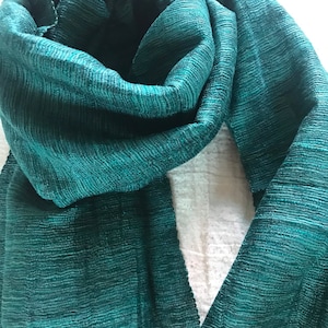 May include: A teal green scarf made of a textured fabric. The scarf is folded in half and draped over a white surface.
