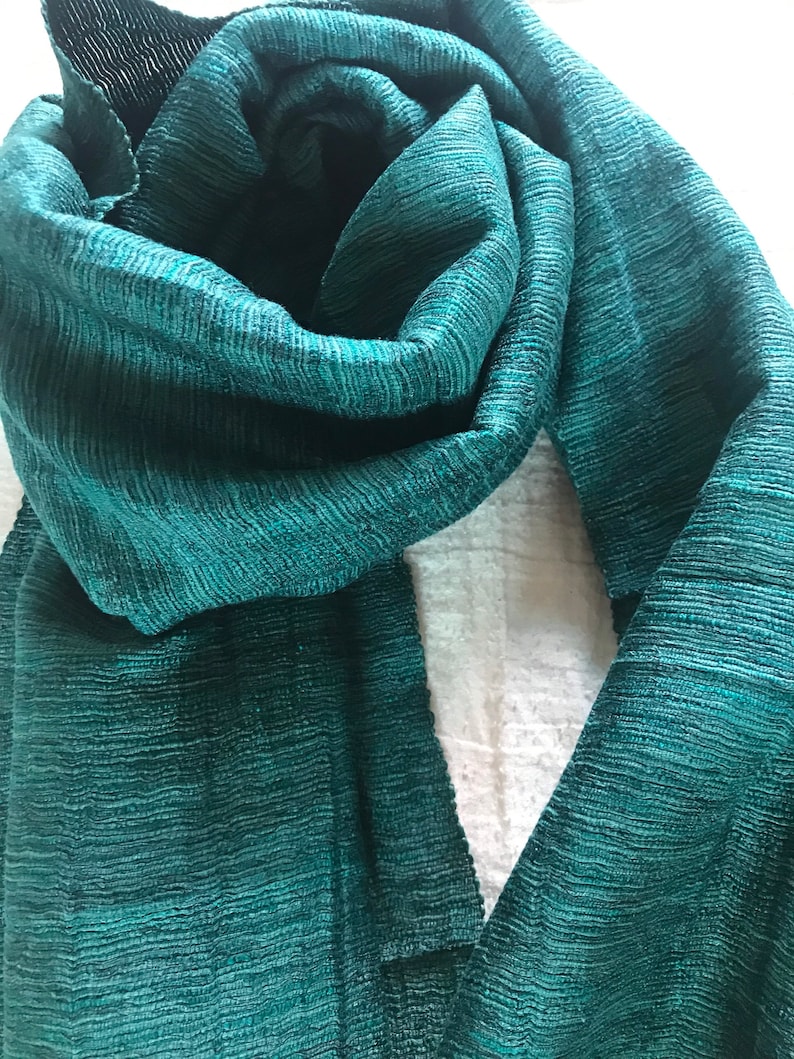 May include: A teal green scarf made of a textured fabric. The scarf is folded in half and draped over a white surface.