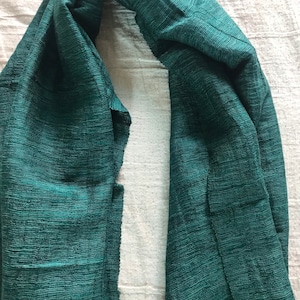 May include: A dark green, textured scarf made of a soft, woven fabric. The scarf is folded in half and draped over a white surface.