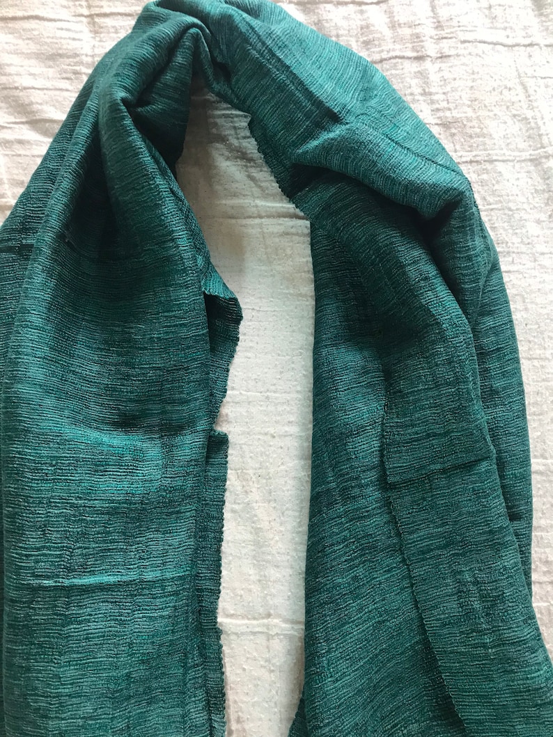May include: A dark green, textured scarf made of a soft, woven fabric. The scarf is folded in half and draped over a white surface.