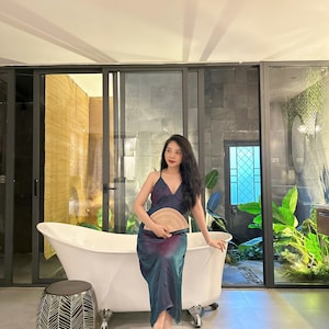 May include: A woman in a blue and purple dress sits on the edge of a white bathtub. She is holding a woven fan in her hands. The bathroom has a large window with a view of a lush green garden.