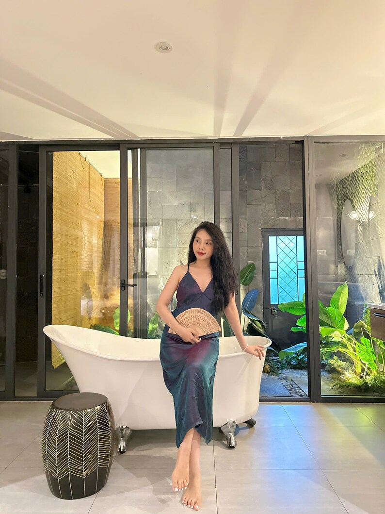 May include: A woman in a blue and purple dress sits on the edge of a white bathtub. She is holding a woven fan in her hands. The bathroom has a large window with a view of a lush green garden.