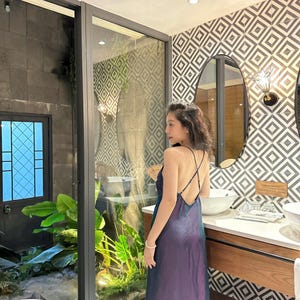 May include: A woman wearing a long, iridescent purple and blue dress with a low back and spaghetti straps. She is standing in a bathroom with a white countertop and a large mirror.