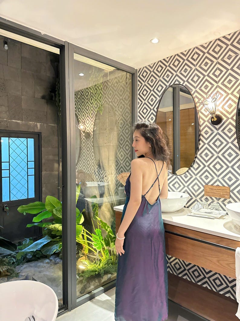 May include: A woman wearing a long, iridescent purple and blue dress with a low back and spaghetti straps. She is standing in a bathroom with a white countertop and a large mirror.