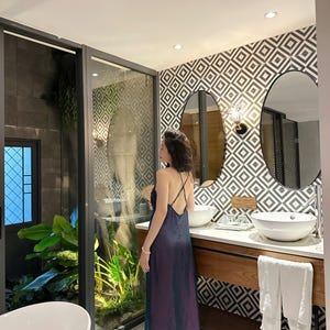May include: A woman in a dark purple dress stands in a bathroom with a white bathtub, a double vanity with two sinks, and a large window with a view of a lush green garden.
