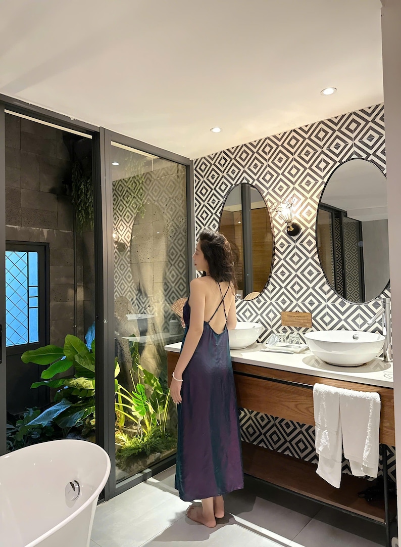 May include: A woman in a dark purple dress stands in a bathroom with a white bathtub, a double vanity with two sinks, and a large window with a view of a lush green garden.