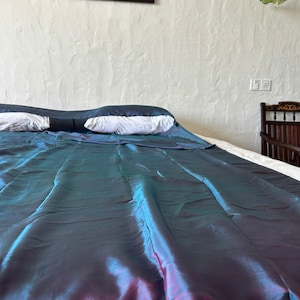 May include: A bed with a teal and purple iridescent bedspread. Two white pillows rest on the bed. The wall behind the bed is white with a textured finish. A framed painting hangs above the bed.