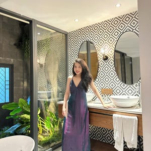 May include: A woman in a purple and blue dress stands in a bathroom with a white sink and a white towel hanging on a rack. The bathroom has a geometric patterned wall and two oval mirrors.