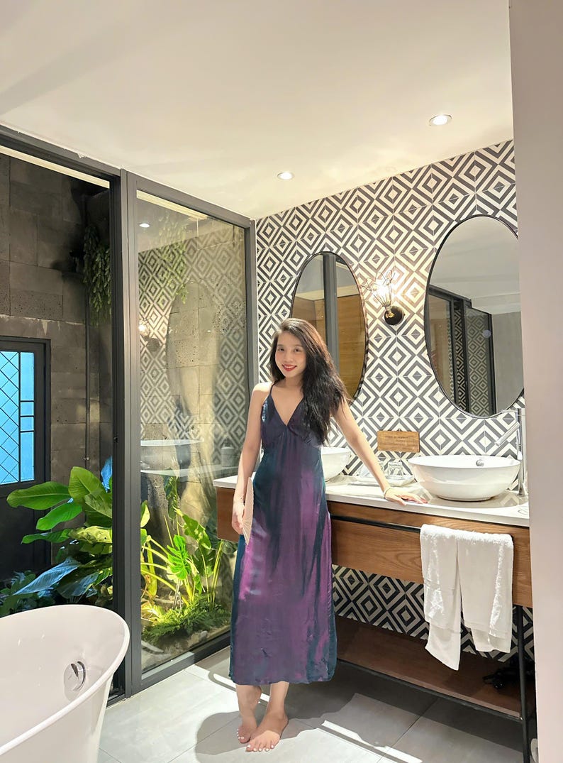 May include: A woman in a purple and blue dress stands in a bathroom with a white sink and a white towel hanging on a rack. The bathroom has a geometric patterned wall and two oval mirrors.