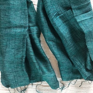 May include: A teal green silk scarf with a textured weave. The scarf is folded in half and the ends are frayed.