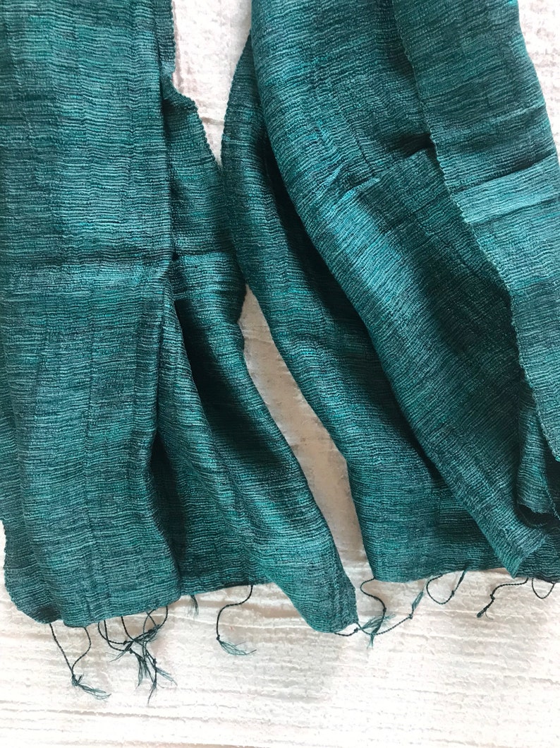 May include: A teal green silk scarf with a textured weave. The scarf is folded in half and the ends are frayed.