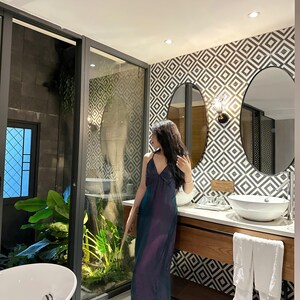 May include: A woman in a long, iridescent blue and purple dress stands in a bathroom with a white sink and a white bathtub. The bathroom has a geometric patterned tile wall and a large window with a view of a lush green garden.