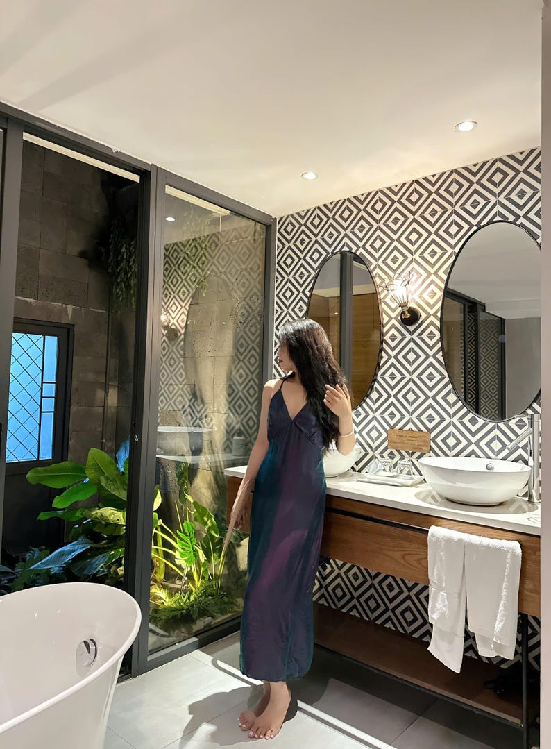 May include: A woman in a long, iridescent blue and purple dress stands in a bathroom with a white sink and a white bathtub. The bathroom has a geometric patterned tile wall and a large window with a view of a lush green garden.