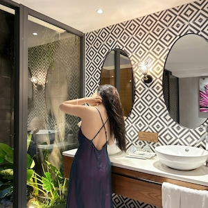 May include: A woman in a dark purple and green satin dress stands in a bathroom with a geometric tiled wall and a double vanity with white sinks.