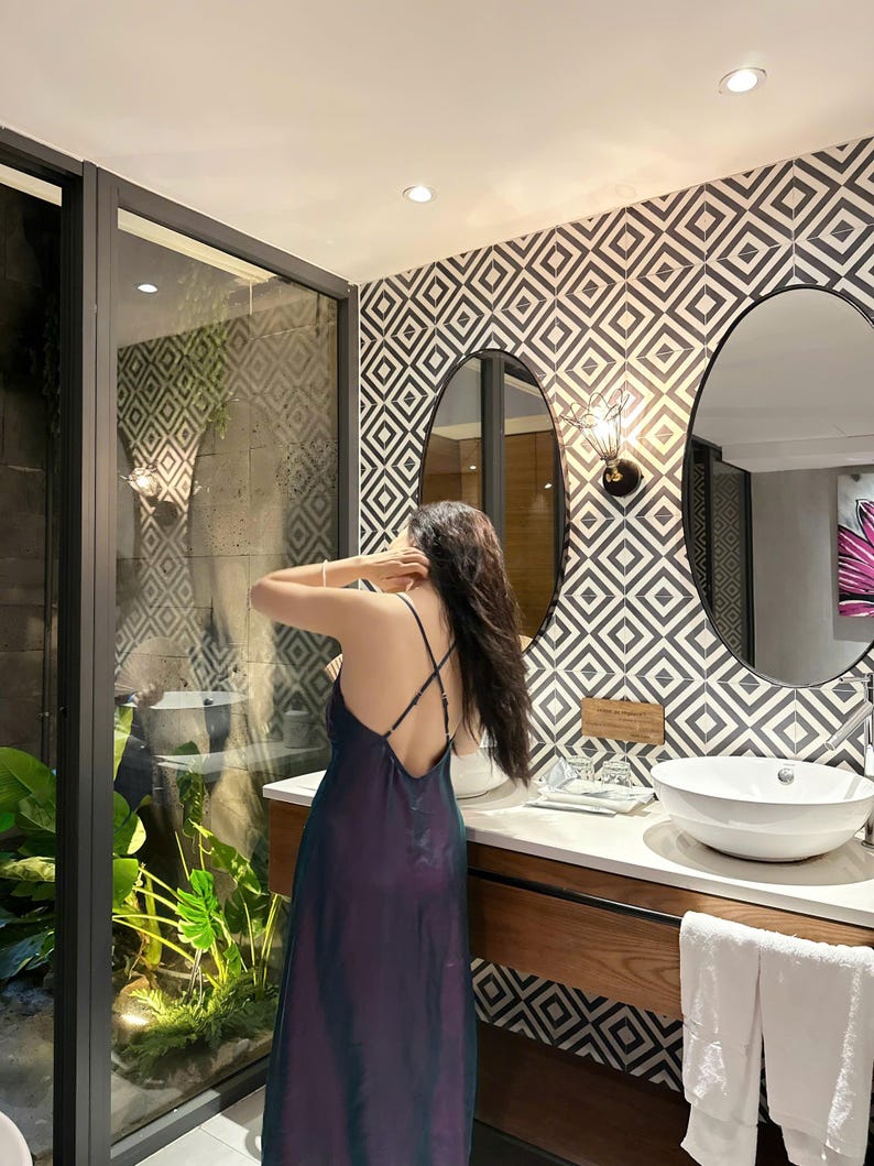 May include: A woman in a dark purple and green satin dress stands in a bathroom with a geometric tiled wall and a double vanity with white sinks.