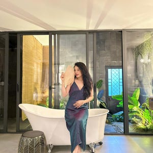 May include: A woman in a blue and purple satin dress sits on the edge of a white bathtub. She is holding a large, round, beige fan in front of her face. The bathroom has a large window with a view of a lush, green garden.