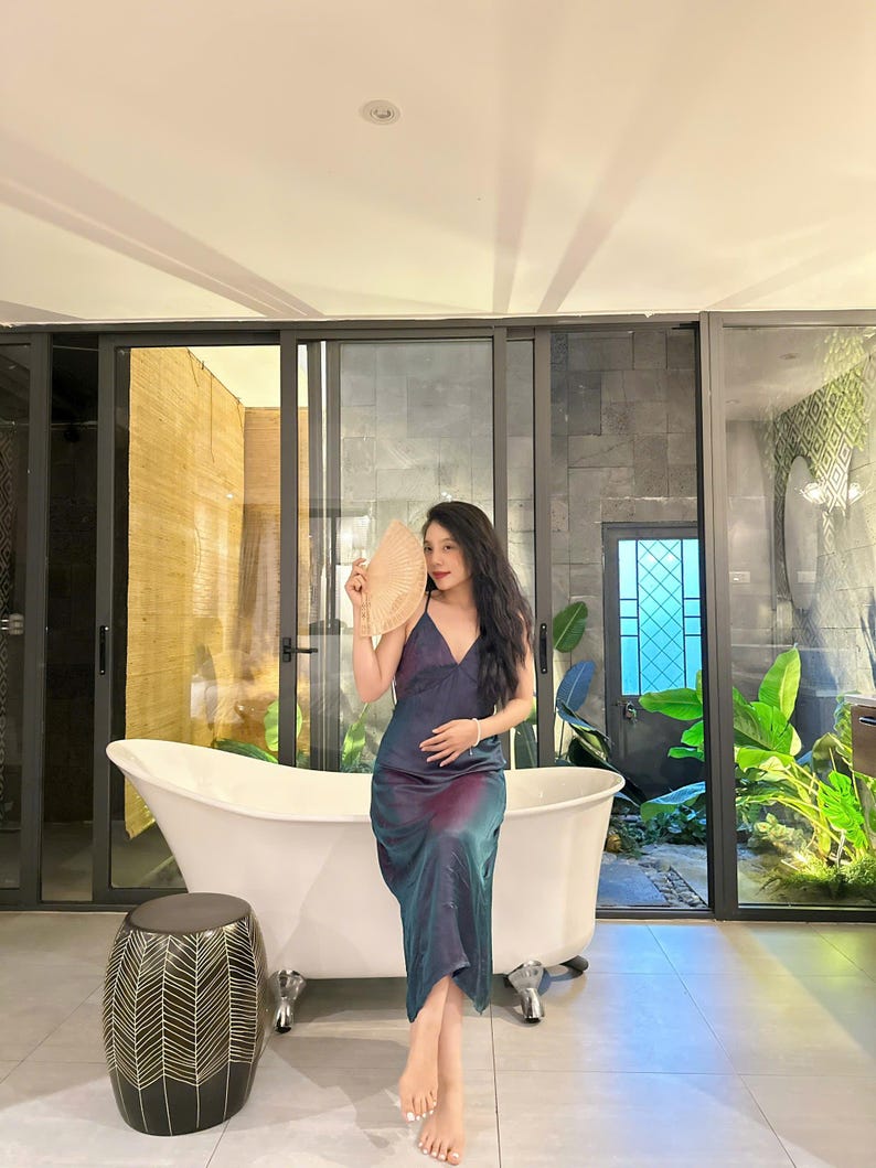 May include: A woman in a blue and purple satin dress sits on the edge of a white bathtub. She is holding a large, round, beige fan in front of her face. The bathroom has a large window with a view of a lush, green garden.