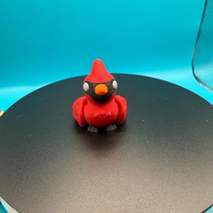 May include: A 3D-printed red cardinal figurine with black accents. The bird has a pointed crest, a black mask around its eyes, and a small orange beak. The figurine is perched on a black surface against a blue backdrop.