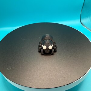 May include: A black 3D-printed spider figurine with white eyes. The spider is positioned on a black circular platform against a blue backdrop. The figurine has a detailed, textured surface.
