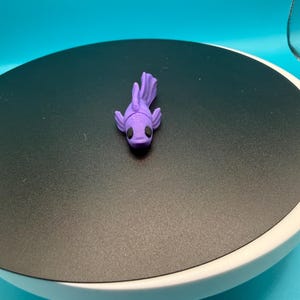 May include: A small, purple, 3D-printed fish figurine with black eyes. The fish is resting on a black surface, set against a blue backdrop. The figurine has a detailed, textured design.