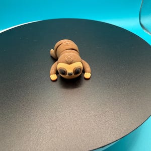 May include: A small, brown sloth figurine with a detailed wood-grain texture. The sloth has a light tan face, large black eyes, and small orange feet. It is lying on a black surface with a blue background.