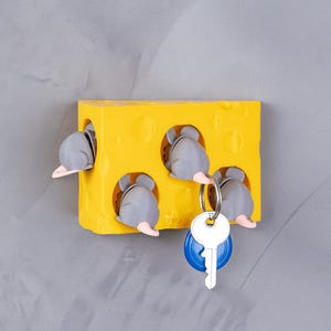 May include: A whimsical yellow cheese-shaped key holder with four gray mice peeking out of the holes. A key with a blue fob hangs from the holder. The key holder is mounted on a gray wall.