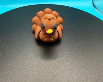 May include: A 3D-printed brown turkey figurine with a yellow beak and red wattle. The turkey has black eyes and a textured surface. The figurine is a Thanksgiving decoration.