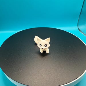 May include: A small, off-white fox figurine with black eyes and a fluffy tail. The fox is standing on a black circular surface against a bright blue background. The figurine is a miniature decorative item.