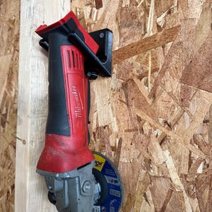 May include: A red and black Milwaukee angle grinder mounted on a wooden surface. The grinder has a silver grinding wheel attached and is secured by a black bracket. The background is a light brown wood.