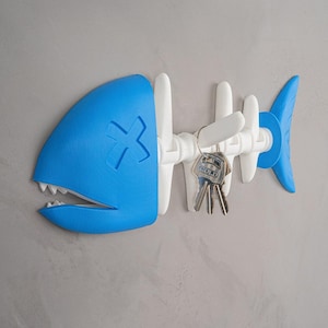 May include: A blue and white fish-shaped key holder with a stylized design. The head is blue with an 'X' marking, and the body is white. Keys hang from the holder, which is mounted on a gray wall. The key holder is a decorative and functional item.