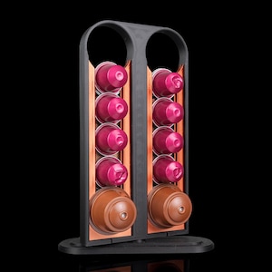 May include: A black plastic coffee pod holder with two vertical columns. Each column holds four small magenta coffee capsules and one larger brown coffee capsule at the base. The frame features a copper-colored accent strip behind the capsules.