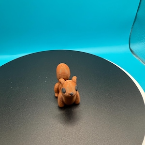May include: A small, 3D-printed squirrel figurine in shades of brown. The squirrel has a textured surface, black eyes, and a bushy tail. The figurine is displayed on a black surface with a blue background.