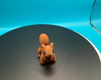May include: A small, 3D-printed squirrel figurine in shades of brown. The squirrel has a textured surface, black eyes, and a bushy tail. The figurine is displayed on a black surface with a blue background.