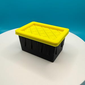 May include: A small, rectangular storage container. It has a black base and a bright yellow lid with a diamond pattern. The container is on a white surface against a teal background.
