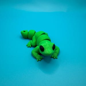 May include: A green plastic toy lizard with black eyes. The lizard is posed in a crawling position with its head raised.