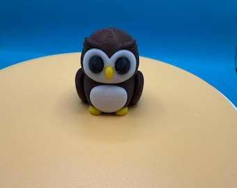 May include: A brown and white owl figurine with large black eyes. The owl is sitting on a light brown surface.