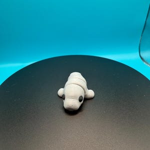 May include: A small, light gray manatee figurine with black eyes and small flipper-like appendages. The figurine is resting on a black surface with a blue background. The manatee is a handmade craft item.