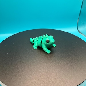 May include: A small, bright green iguana figurine with a segmented body and spiky dorsal ridge. The iguana has small, detailed feet and a round head with black eyes. It is displayed on a black circular surface against a teal background.