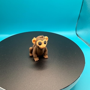 May include: A brown and tan 3D-printed monkey figurine with large, round eyes and a smiling expression. The monkey is in a crouching position on a black surface, set against a bright blue background. The figurine is a detailed representation of a monkey.