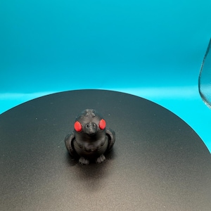 May include: A small, black 3D-printed bird figurine with bright red eyes. The bird is sitting on a black surface against a bright blue background. The figurine has a textured surface and a rounded body.