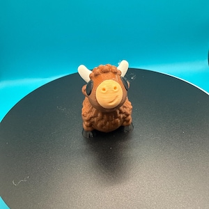 May include: A 3D-printed figurine of a Highland cow. The cow has a brown body, a tan face, black eyes, and white horns. The figurine is sitting on a black surface against a teal background.