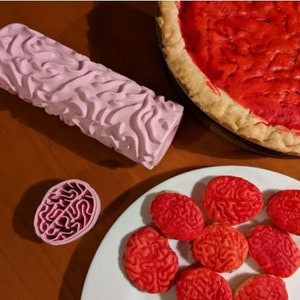 May include: A pink brain-shaped rolling pin and a plate of red brain-shaped cookies. The cookies are on a white plate with a blue background.
