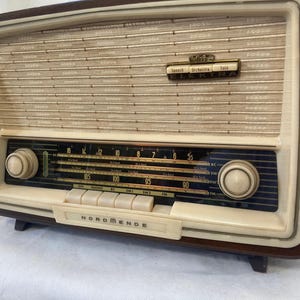 Puede incluir: Radio Nordmende Elektra vintage en crema y marrón. Cuenta con una rejilla de altavoz, un dial de sintonización y botones. La radio rectangular está hecha de madera y plástico. La palabra 'Nordmende' está impresa en la parte delantera.
